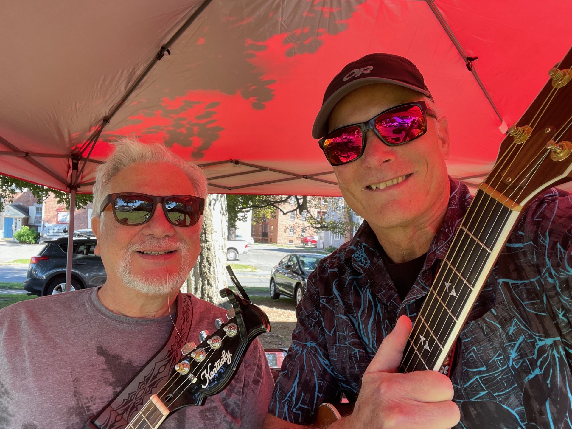 Two men wearing sunglasses, smiling and holding guitars.